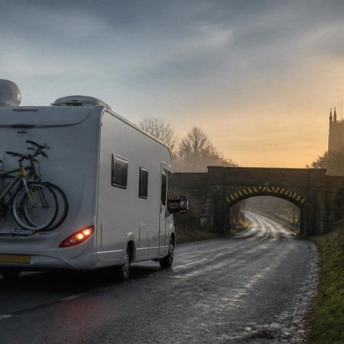 Coachbuilt motorhome with a satellite dome, air conditioning unit and bikes on the rack approaching a low railway bridge at dawn